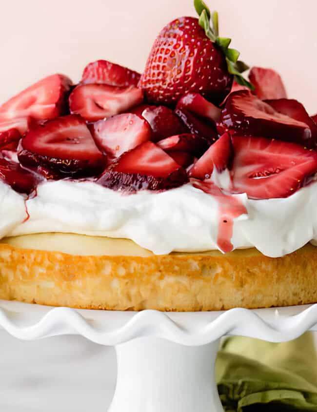 A strawberry shortcake topped with whipped cream and fresh sliced strawberries sits on a white, ruffled cake stand. The background is a soft pink, and a small green cloth is partially visible at the base of the cake stand. White flowers are placed next to this delightful example of strawberry desserts.