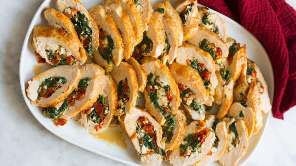 A white platter filled with sliced stuffed chicken breasts, perfect for any collection of Chicken Recipes. The chicken appears tender and juicy, with each slice revealing a colorful filling of spinach, sun-dried tomatoes, and crumbled feta cheese. The platter is set on a marble surface.