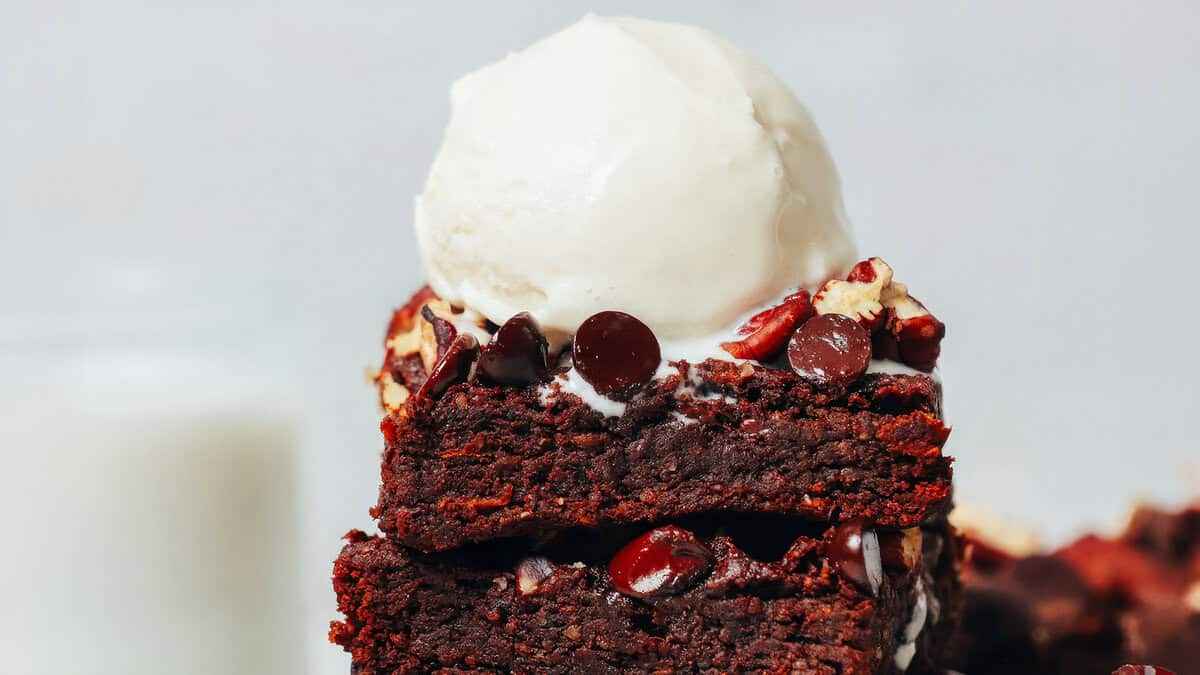 A close-up of stacked fudgy brownies topped with chocolate chips and nuts, crowned with a scoop of vanilla ice cream. Perfect for chocoholics seeking a treat that goes beyond basic, the background features a blurred glass of milk.