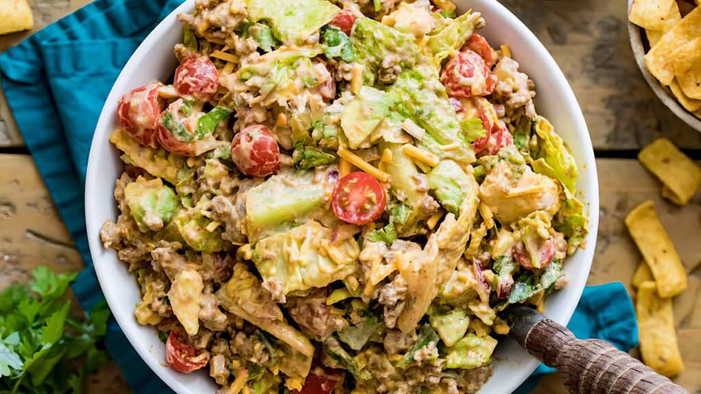 A vibrant bowl of taco salad filled with fresh ingredients like chopped lettuce, cherry tomatoes, shredded cheese, ground beef from tried-and-true Ground Beef Recipes, and avocado slices. The salad is dressed with a creamy, slightly spicy dressing. Corn chips are visible on the side.