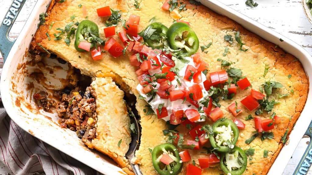 A baked casserole dish filled with tamale pie is topped with sliced jalapeños, chopped tomatoes, fresh cilantro, and a dollop of sour cream. A large portion has been served, revealing a filling of ground beef, beans, and vegetables beneath the cornbread crust—perfect for fans of ground beef recipes.