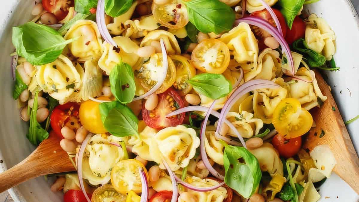 A vibrant salad for pasta lovers, featuring tortellini pasta, halved cherry tomatoes (red and yellow), white beans, thinly sliced red onions, and fresh basil leaves. The ingredients are tossed together in a white bowl with wooden serving utensils.