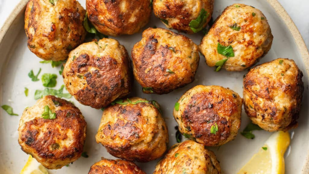 A plate of golden-brown meatballs garnished with chopped parsley, arranged in a close-up view, reminiscent of a featured dish from an enticing cookbook. The meatballs appear crispy and well-seasoned, with a wedge of lemon positioned near the edge of the plate.