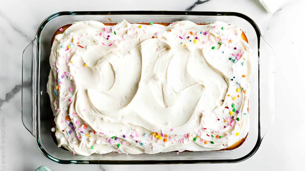 A rectangular cake in a glass dish, topped with white buttercream frosting and colorful sprinkles. The frosting is spread with gentle, swirling patterns, and the edges of the cake are visible slightly through the glass sides of the dish. Perfect for those searching for delightful sheet cake recipes.