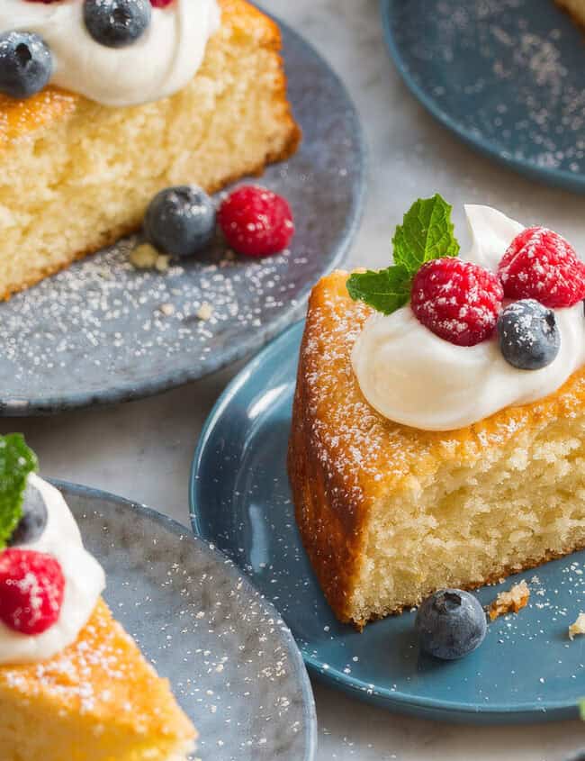 Slices of sponge cake garnished with dollops of whipped cream, fresh blueberries, and raspberries—classic fruit desserts—are served on blue plates. A blue cloth napkin and a silver fork are also visible, with a light dusting of powdered sugar on the marble countertop.