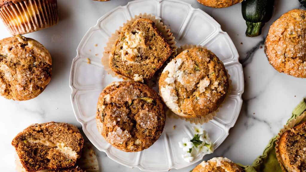 A white plate holds three zucchini muffins with sugar-dusted tops, one muffin is cut in half to reveal its crumbly interior. Surrounding the plate are more muffins on a marble surface, along with a small piece of zucchini and flower garnishes, perfect for those exploring new muffin recipes.