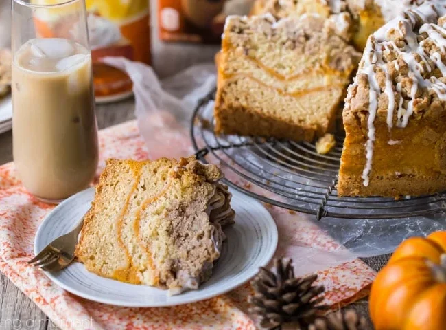 A slice of cake sits on a white plate with a fork beside it. Behind the plate, there is a wire rack holding a large, partly sliced coffee cake drizzled with icing. An iced coffee, small decorative pumpkins, and pine cones are also visible on a table with an orange-patterned cloth.