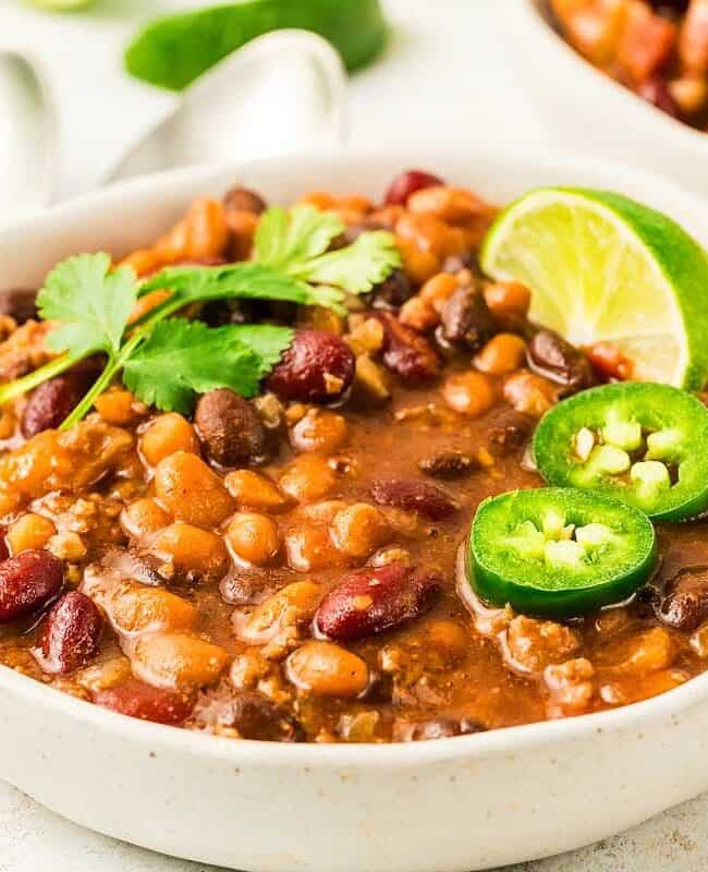 A bowl of hearty chili filled with beans, ground meat, and rich tomato sauce, garnished with cilantro, sliced jalapenos, and a wedge of lime—perfect for slow cooker recipes. A second bowl and spoons are blurred in the background. Fresh cilantro is also visible nearby.