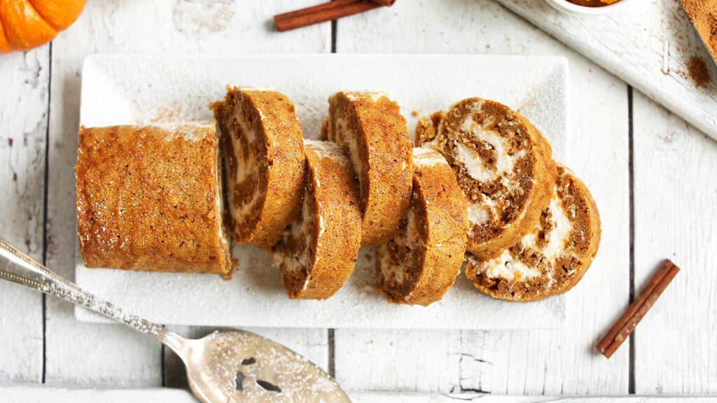 A sliced pumpkin roll with cream cheese filling is displayed on a rectangular white plate. The table underneath is light wood, with a silver serving spatula and two cinnamon sticks placed nearby, giving the scene a festive feel.