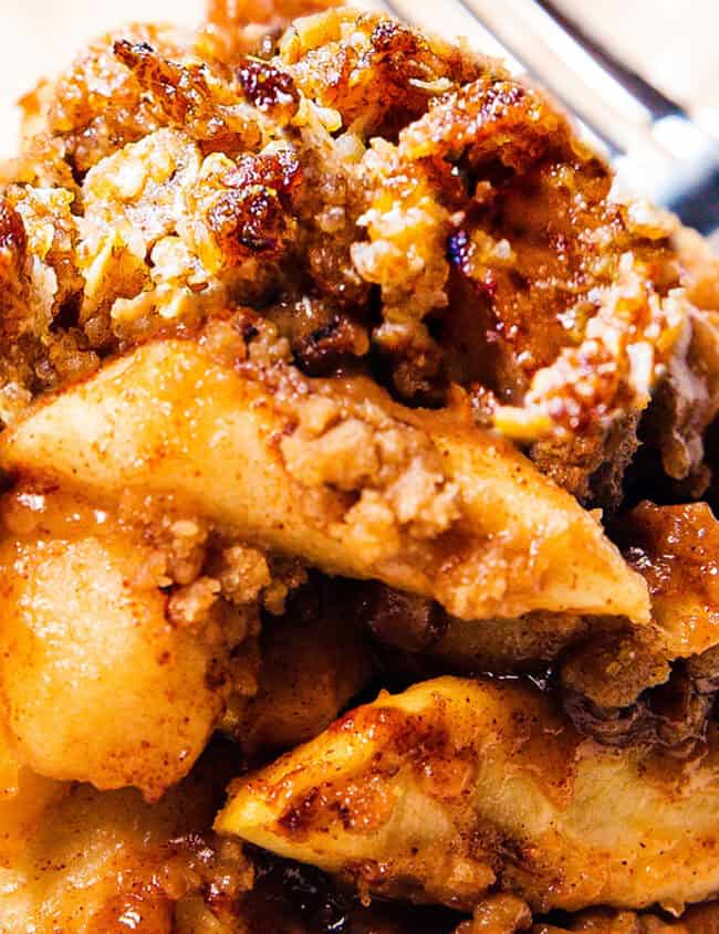 Close-up of a serving of apple crisp, featuring baked apple slices topped with a golden, crispy oat and sugar mixture. The dessert is swimming in a caramel-like sauce, delivering those delicious fall flavors. A fork is placed to the right, partially visible on a white plate—a must-try for apple recipes lovers.