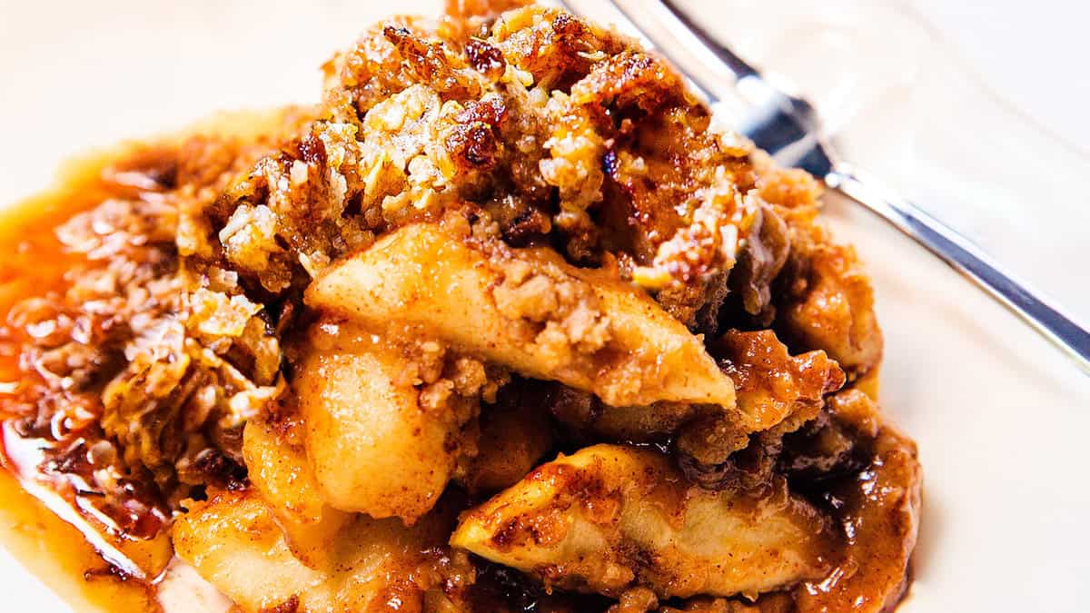 Close-up of a serving of apple crisp, featuring baked apple slices topped with a golden, crispy oat and sugar mixture. The dessert is swimming in a caramel-like sauce, delivering those delicious fall flavors. A fork is placed to the right, partially visible on a white plate—a must-try for apple recipes lovers.