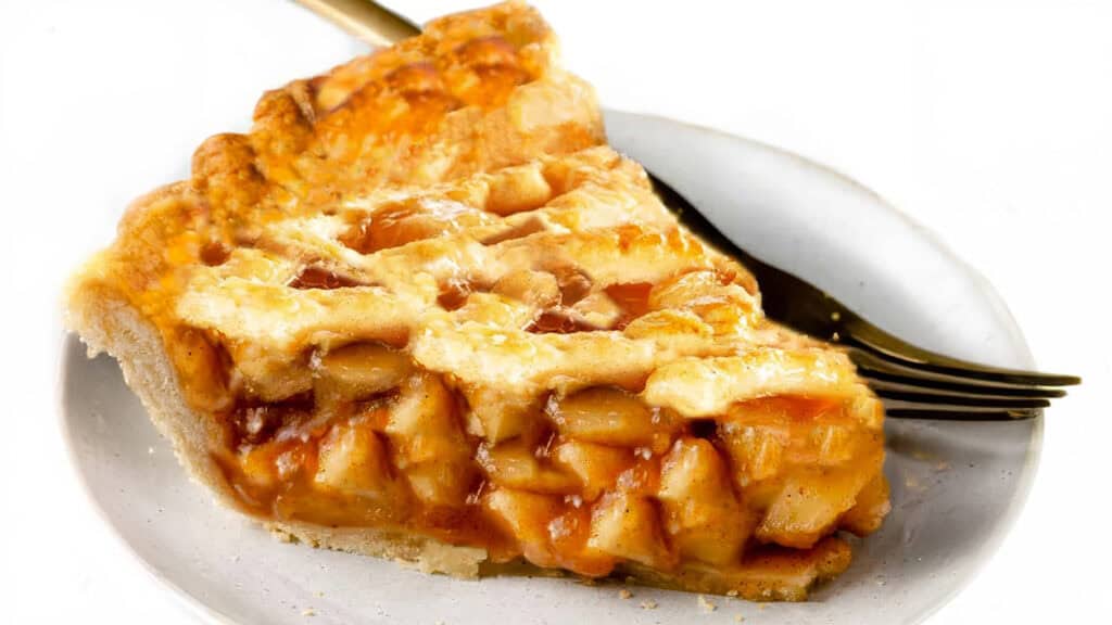 A slice of apple pie with a flaky, lattice top crust is placed on a white plate. A gold fork is beside the pie, resting on the plate. The gooey, caramelized filling features chunks of apples, embodying fall flavors and delicious recipes at their finest.