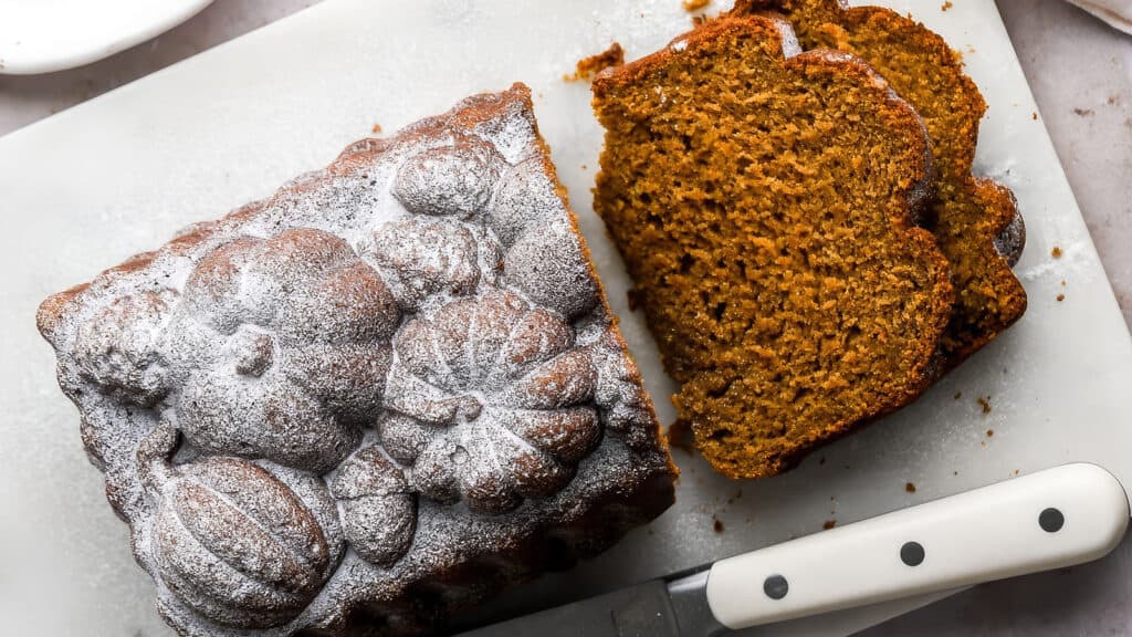 A loaf of pumpkin bread topped with powdered sugar, decorated with pumpkin shapes, placed on a white cutting board. Two slices have been cut from the loaf and lie next to it. A knife with a white handle is also on the board.