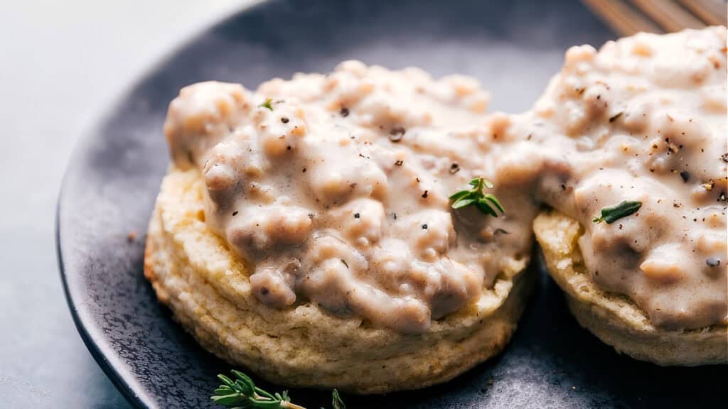 A close-up of two Southern biscuits topped with sausage gravy. The gravy is speckled with ground pepper and small bits of sausage. Fresh thyme leaves garnish the dish, which is served on a dark plate in a well-lit setting.