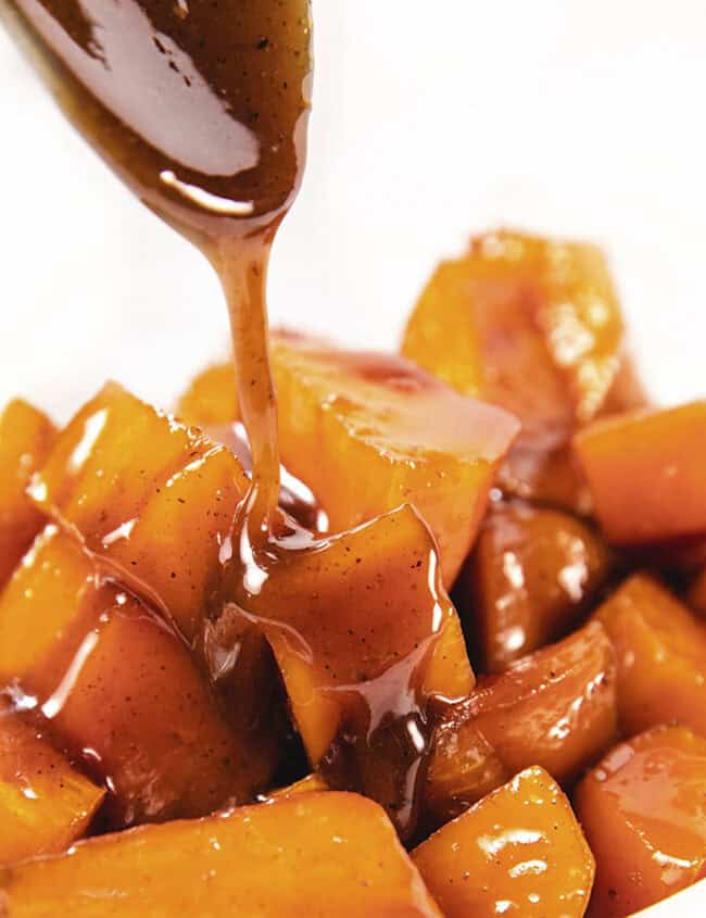 A bowl filled with Candied Sweet Potatoes, glistening with a brown, glossy sauce. A spoon is seen drizzling more of the rich caramel sauce over the Candied Sweet Potatoes, evoking classic Southern recipes and creating an inviting appearance.