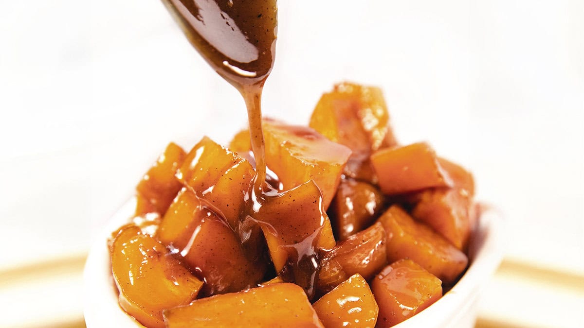 A bowl filled with Candied Sweet Potatoes, glistening with a brown, glossy sauce. A spoon is seen drizzling more of the rich caramel sauce over the Candied Sweet Potatoes, evoking classic Southern recipes and creating an inviting appearance. 