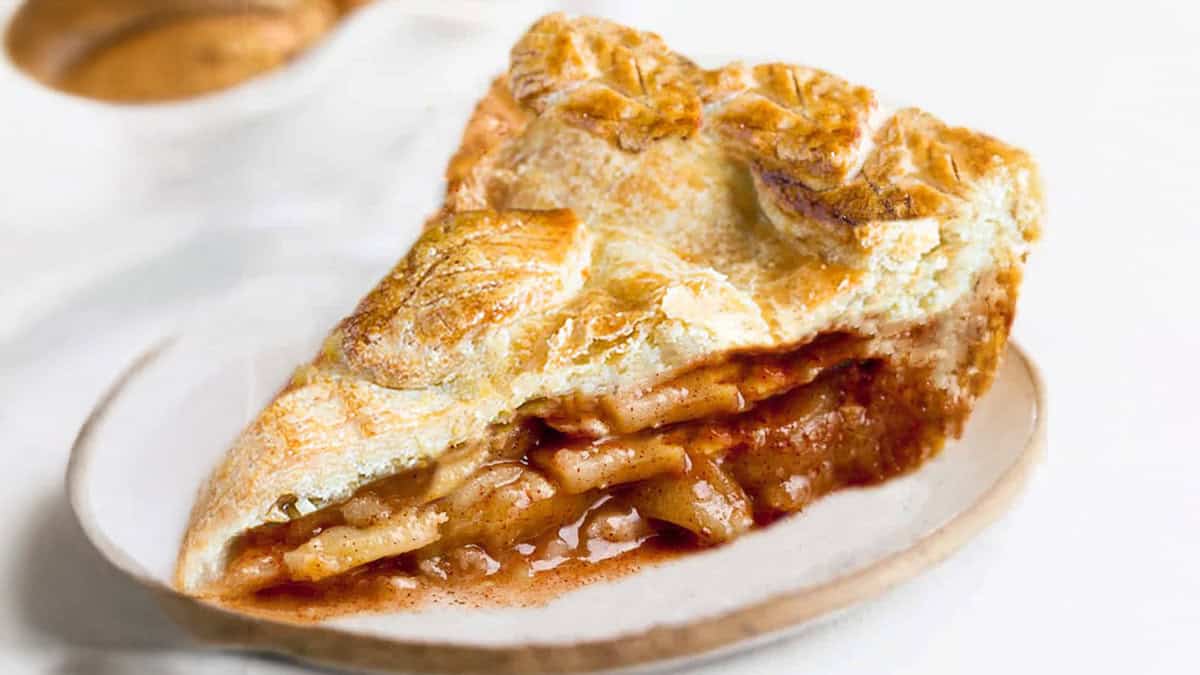 A close-up shot of a slice of apple pie on a white plate. The pie has a golden-brown, flaky crust with visible layers of baked apple slices and cinnamon, embodying the essence of Thanksgiving desserts. A small portion of sauce is oozing out from the filling, and the background is softly blurred.