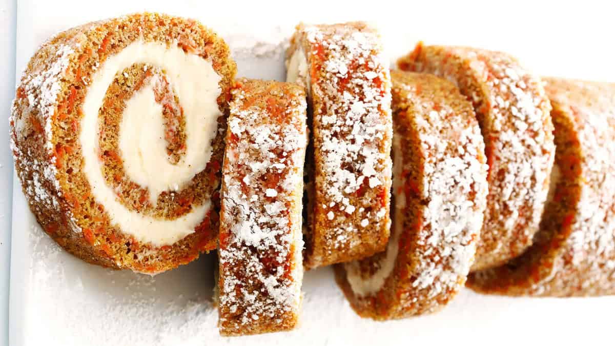 A close-up of a sliced cake roll filled with cream cheese frosting. The lightly browned roll has a swirl pattern and is dusted with powdered sugar. The inner cream layer contrasts with the slightly crusty outer layer. The slices are neatly arranged in a line.