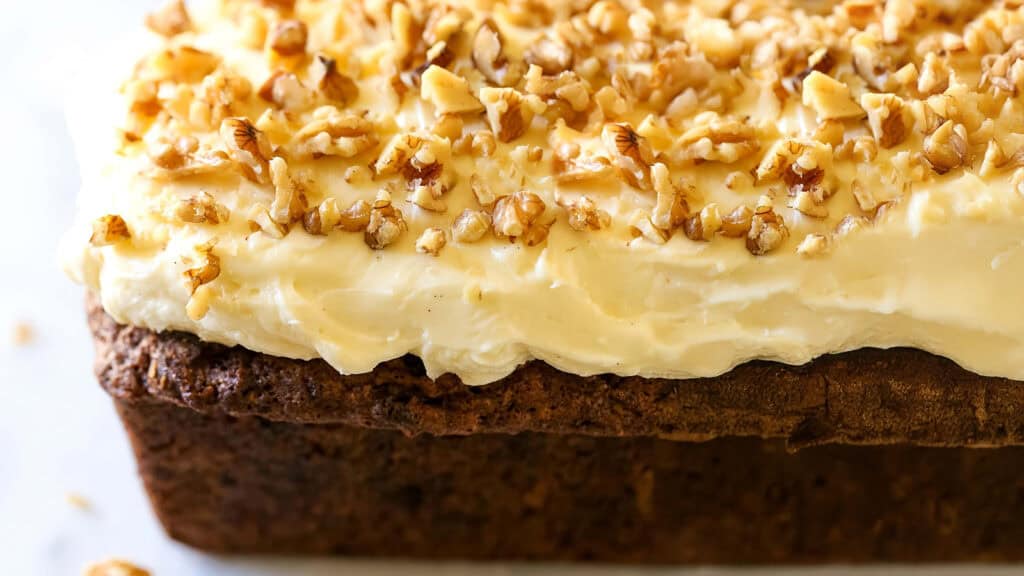 A close-up of a loaf of carrot cake topped with a thick layer of creamy frosting and a generous sprinkle of chopped walnuts. Perfect for desserts, this bakery delight appears moist and dense, with bits of carrots visible, and the frosting looks smooth and rich.