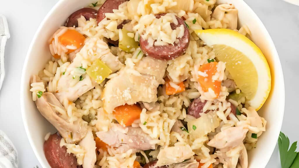         A bowl of chicken and sausage jambalaya, filled with rice, sliced sausages, chunks of chicken, chopped carrots, celery, and a lemon wedge garnish. The ingredients are mixed and cooked together, showcasing a hearty and colorful Southern recipe.