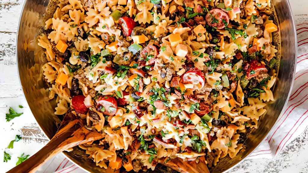 A large bowl of vibrant pasta salad with bowtie pasta, cherry tomatoes, diced cheese, black olives, and garnished with fresh chopped herbs sits elegantly. Wooden salad tongs rest in the bowl on a striped cloth. For a twist on ground beef recipes, consider adding savory crumbles to this dish.