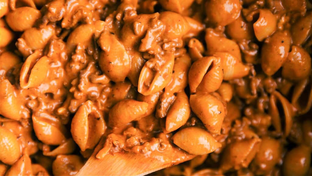 Close-up of creamy, cheesy pasta shells mixed with ground beef. The pasta is coated in a rich, orange-colored sauce, creating a hearty and comforting dish. A wooden spoon is lifting a portion from the pan, showcasing one of the best ground beef recipes you'll ever try.