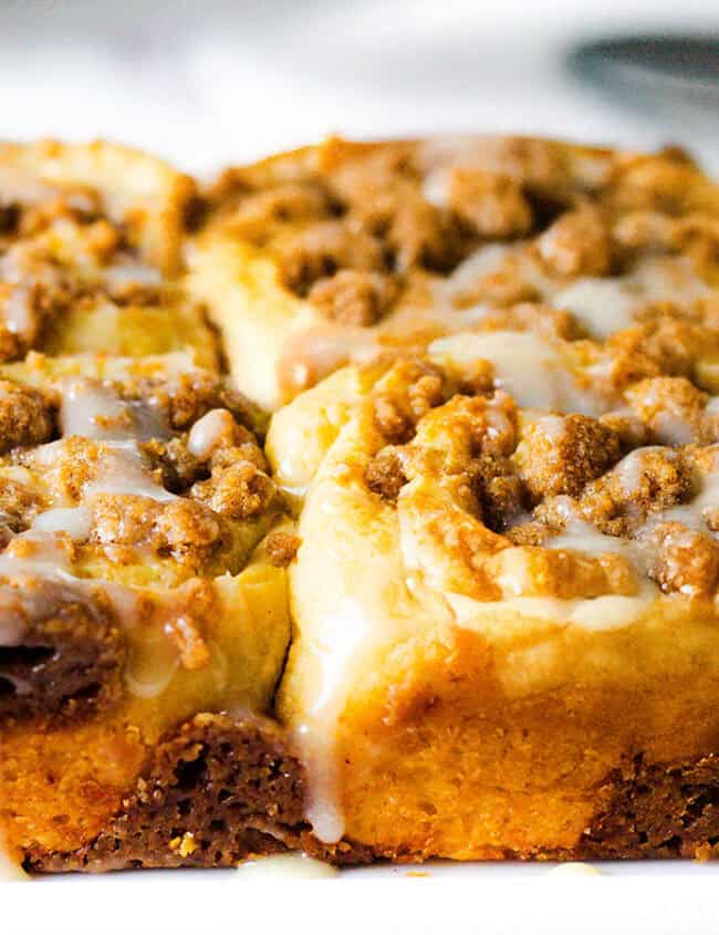 A close-up of a batch of freshly baked crumb-topped coffee cake squares, drizzled with a light glaze. The golden-brown cake is topped with a crumbly, cinnamon-infused streusel, and the glaze slightly overflows, adding a sweet finish to this quintessential fall dessert.