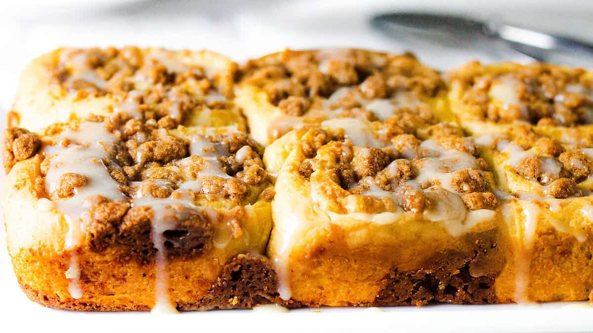 A close-up of a batch of freshly baked crumb-topped coffee cake squares, drizzled with a light glaze. The golden-brown cake is topped with a crumbly, cinnamon-infused streusel, and the glaze slightly overflows, adding a sweet finish to this quintessential fall dessert.