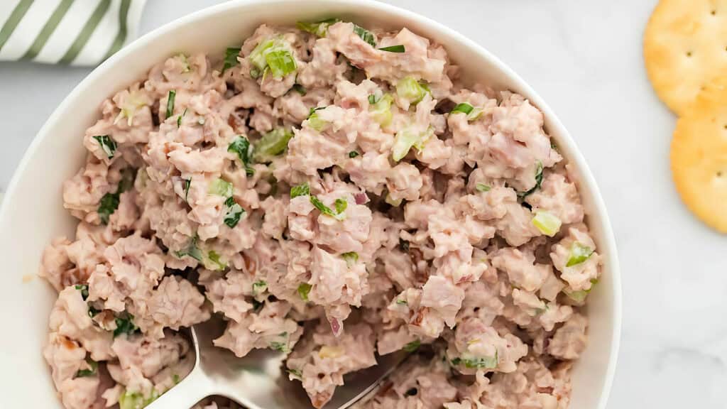 A bowl of chicken salad mixed with chopped celery and herbs, inspired by Southern recipes, is placed on a white surface. A serving spoon rests in the bowl, while two round crackers are partially visible in the upper right corner. A striped cloth can be seen in the top left.