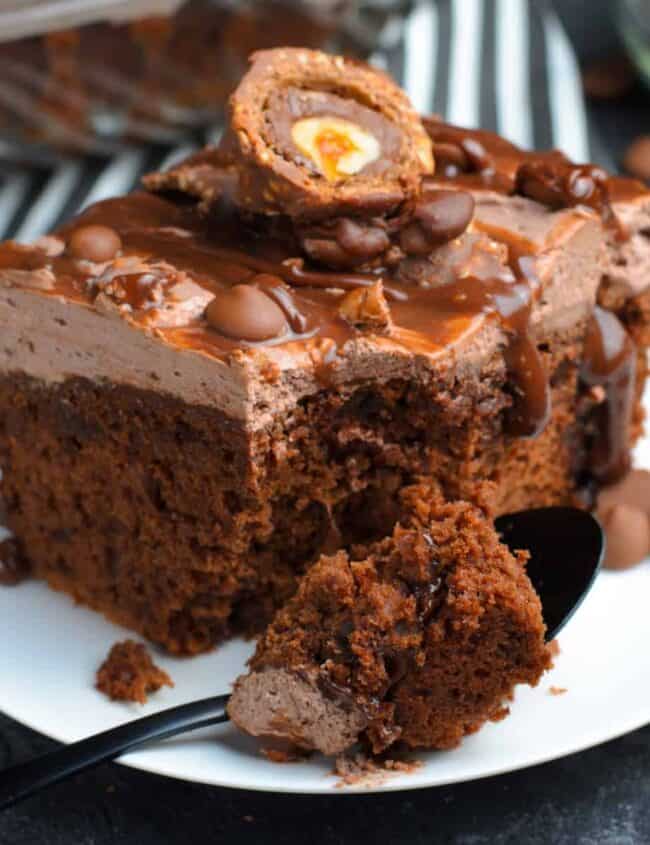 A close-up of a decadent chocolate cake slice on a white plate, garnished with chocolate frosting, chocolate chips, and topped with a halved chocolate egg candy. A black spoon beside the cake holds a small piece. The background features a striped cloth—perfect inspiration for sheet cake recipes.