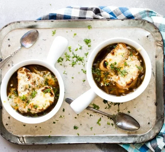 Two bowls of French onion soup, straight from our favorite soup recipes, are topped with melted cheese and garnished with chopped parsley, placed on a rustic metal tray. Each bowl has a spoon beside it, and a blue and white checkered cloth is draped around the tray.