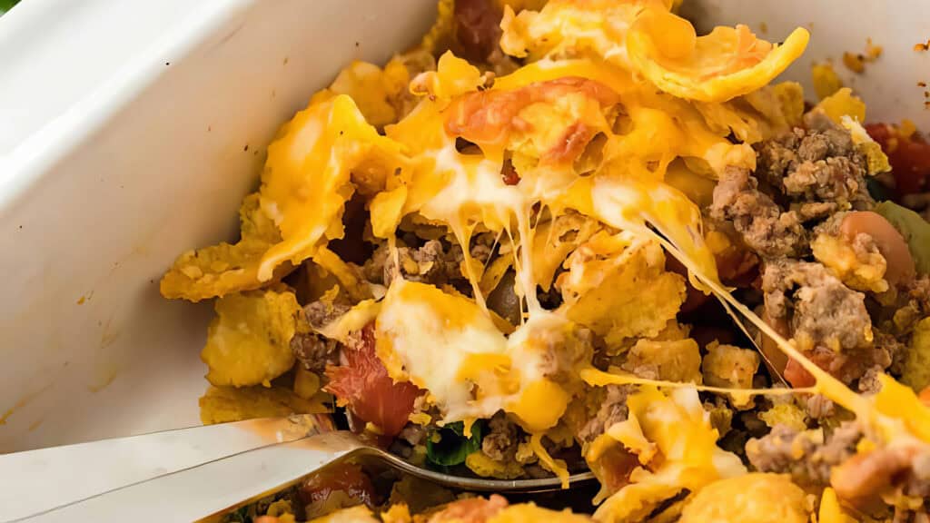 A close-up shot of a loaded nachos dish in a white container. The nachos, inspired by popular ground beef recipes, are topped with melted cheese, ground beef, diced tomatoes, and other toppings. A metal spoon is partially visible, suggesting the dish is being served.