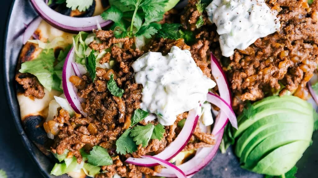 A close-up of a delicious meal featuring seasoned ground beef recipes topped with dollops of creamy sauce. The dish is garnished with cilantro, sliced red onions, and avocado slices, served on a flatbread base, all arranged on a plate.