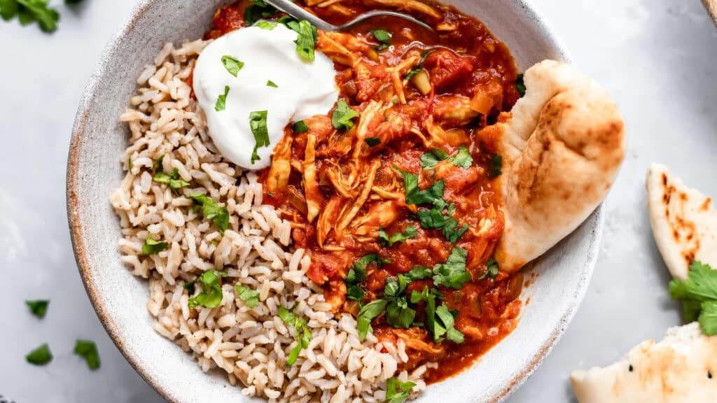 A bowl containing shredded chicken in a rich tomato sauce, garnished with chopped cilantro and a dollop of sour cream on top. On one side, there is a serving of brown rice, and a piece of naan bread is placed on the edge of the bowl—a delightful twist on slow cooker recipes.