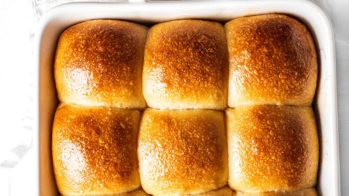 A close-up view of a white rectangular baking dish containing six golden-brown, freshly baked dinner rolls. The tops of the rolls are shiny and evenly browned, suggesting a soft and fluffy texture inside—perfect Thanksgiving sides to complement your festive meal.