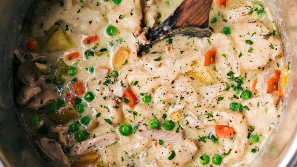 A pot filled with creamy Southern chicken and dumplings. The dish includes chunks of chicken, green peas, diced carrots, and potato pieces, all blended in a thick, creamy broth. Finely chopped parsley is sprinkled on top, and a wooden spoon is stirring the mixture.