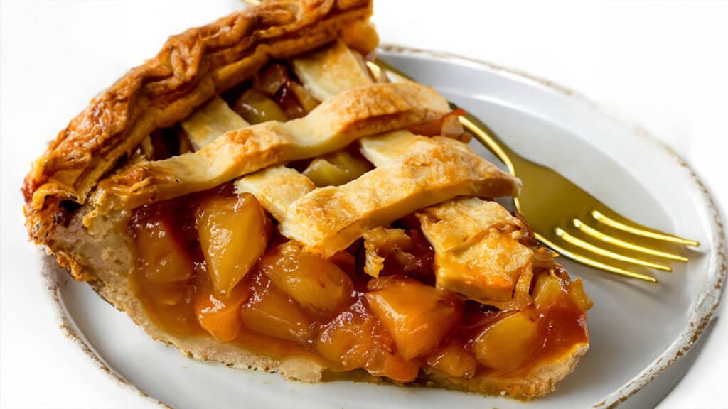 A close-up of a slice of peach pie, showcasing golden-brown lattice crust and juicy peach filling, served on a white plate with a gold-colored fork.