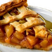 A close-up of a slice of peach pie, showcasing golden-brown lattice crust and juicy peach filling, served on a white plate with a gold-colored fork.