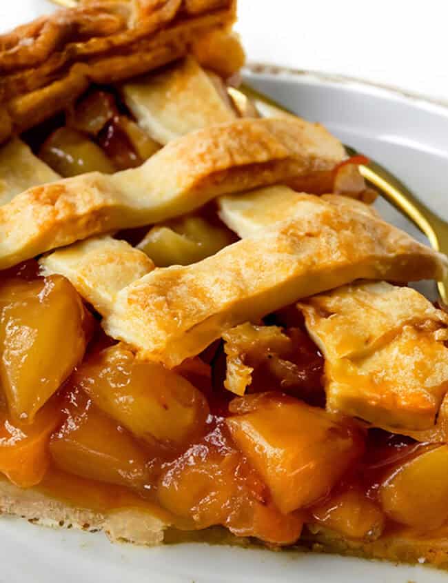 A close-up of a slice of peach pie, showcasing golden-brown lattice crust and juicy peach filling, served on a white plate with a gold-colored fork.