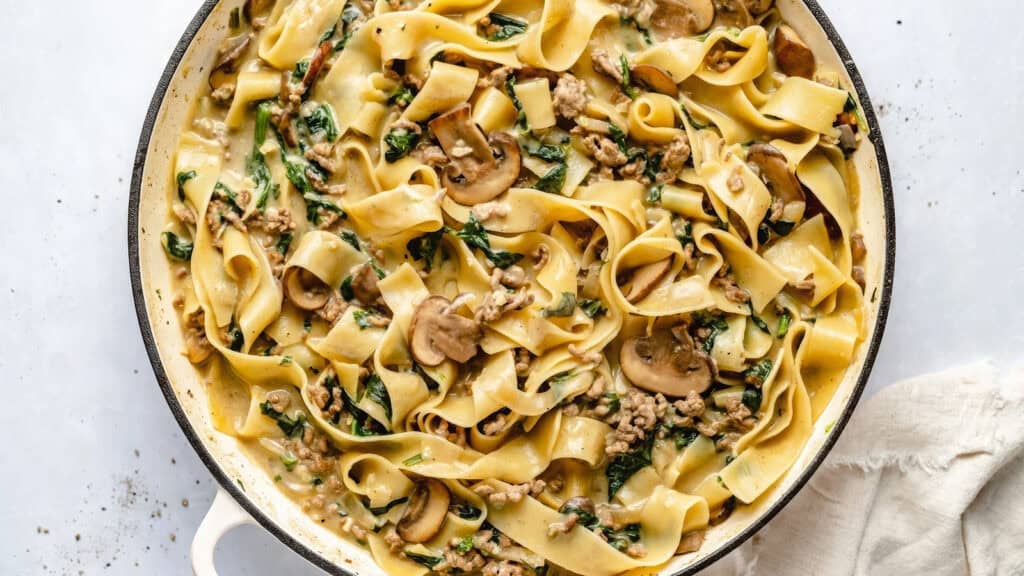 A skillet filled with creamy pappardelle pasta featuring sliced mushrooms, spinach, and ground beef. The wide noodles are coated in a rich, light-colored sauce, and the dish is garnished with bits of herbs and seasonings. A white napkin lies beside the skillet—a standout among ground beef recipes.