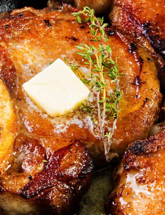 A close-up view of a skillet containing juicy, seared pork chops—a classic Southern comfort food. Each chop is garnished with a sprig of fresh thyme, and the center chop has a melting pat of butter on top. The edges are browned, and whole garlic cloves are scattered around.