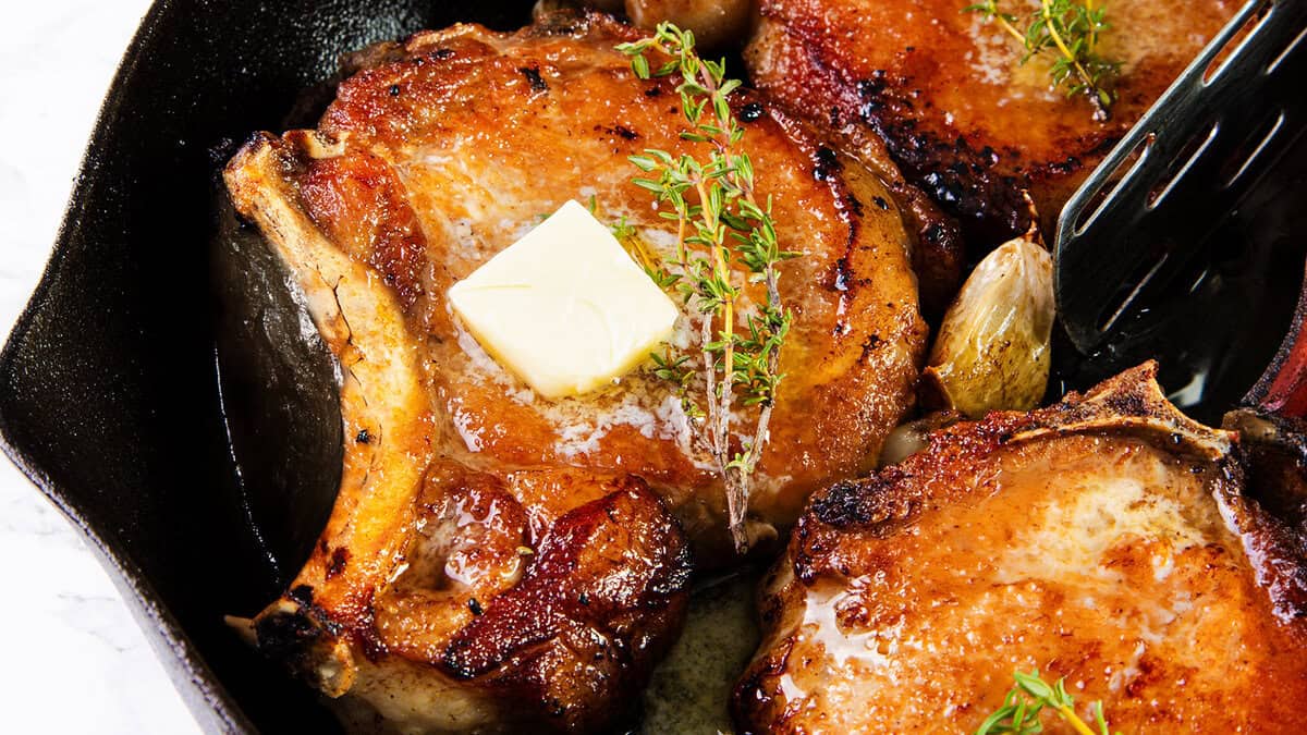 A close-up view of a skillet containing juicy, seared pork chops—a classic Southern comfort food. Each chop is garnished with a sprig of fresh thyme, and the center chop has a melting pat of butter on top. The edges are browned, and whole garlic cloves are scattered around.