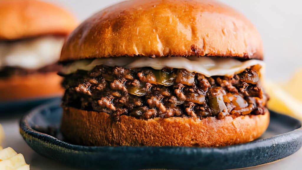 A close-up of a juicy sloppy joe sandwich filled with seasoned ground beef and onions, topped with melted cheese, in a glossy, toasted bun. The sandwich is on a blue plate, with a blurred view of another mouthwatering ground beef recipe in the background.