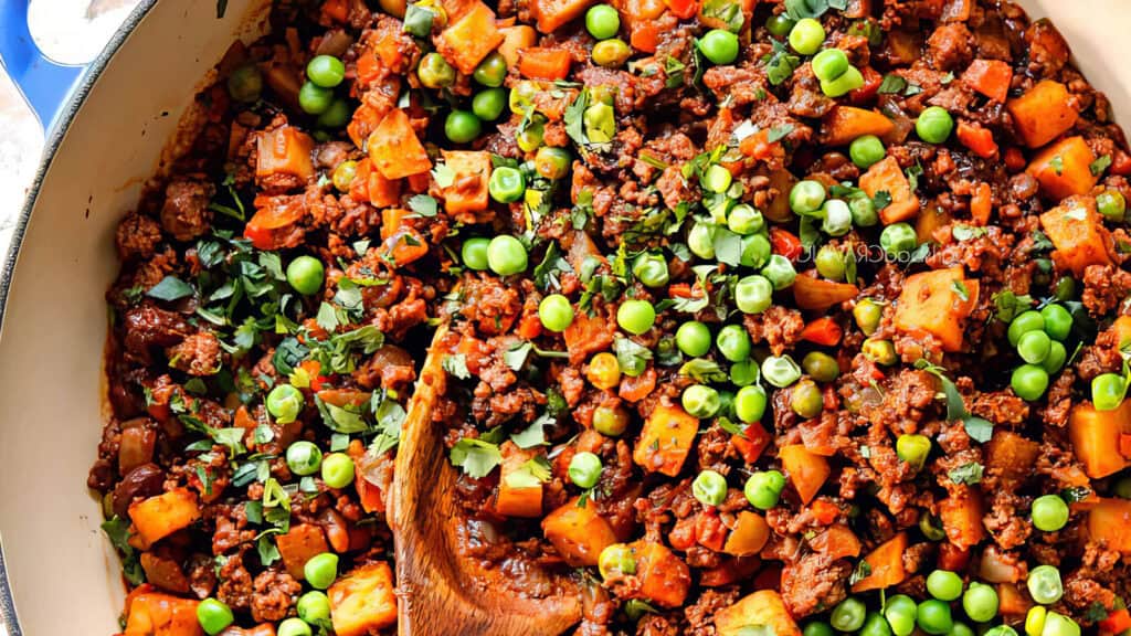 A close-up of a dish featuring ground beef recipes with minced meat mixed with diced vegetables, including carrots, potatoes, and green peas. The dish is garnished with chopped herbs and is being stirred with a wooden spoon in a large pan.