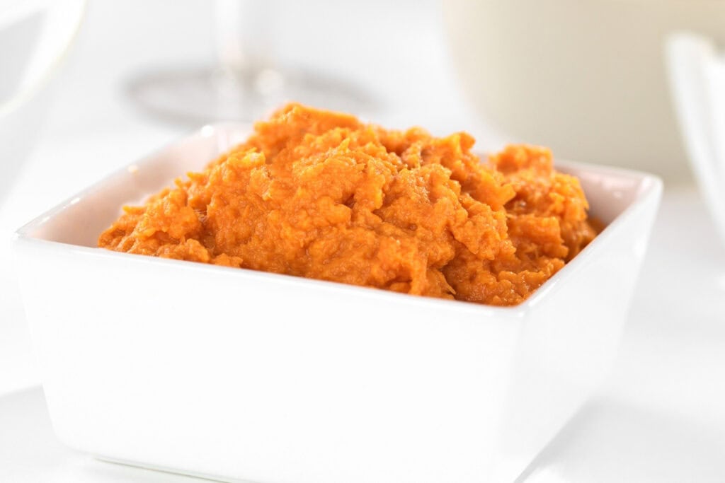 A square white bowl filled with mashed sweet potatoes sits on a white surface, reminiscent of the vibrant hues found in a Pumpkin Swiss Roll. The sweet potatoes are smooth and creamy with a bright orange color. The background is softly blurred, making the dish the focal point.