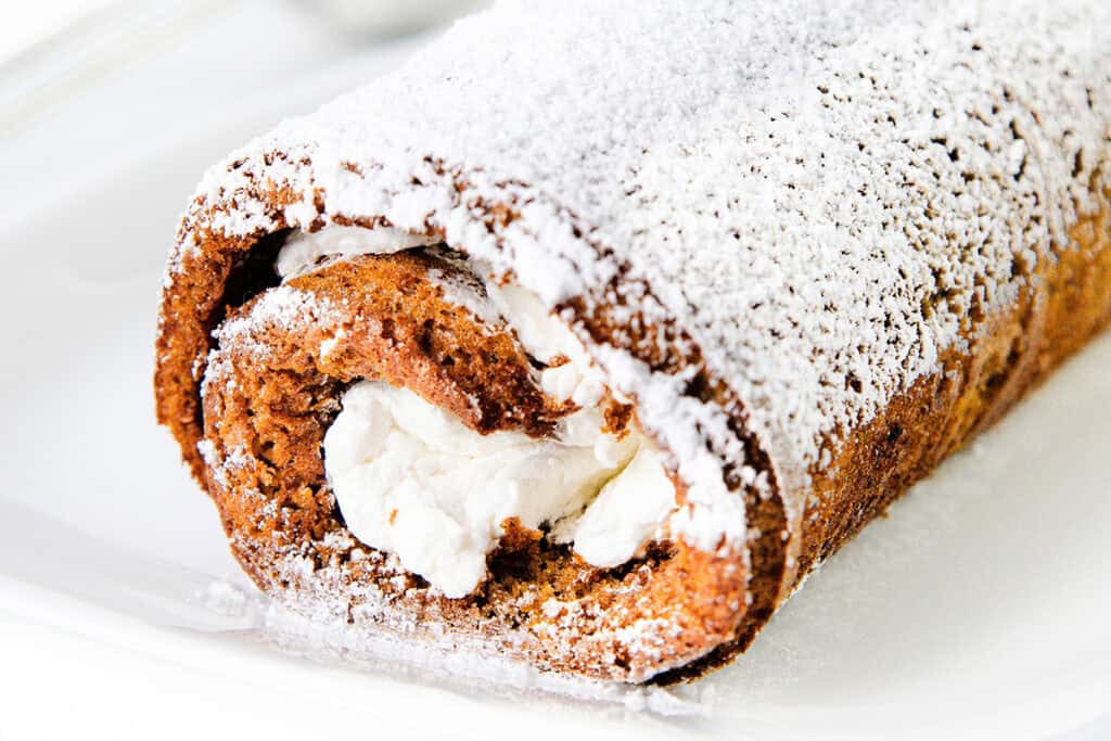 A close-up image of a sliced Pumpkin Swiss Roll cake dusted with powdered sugar. The cake is rolled with a creamy white filling visible in the center, and the outer cake layer appears to be a rich brown color, suggesting a chocolate or spice-flavored cake.