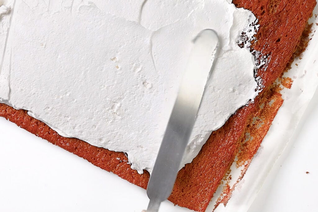 A close-up of a rectangular cake base being spread with white frosting using an offset spatula. The partially frosted edges reveal it's a Pumpkin Swiss Roll, with uncoated sides and the cake resting on parchment paper or a similar surface.