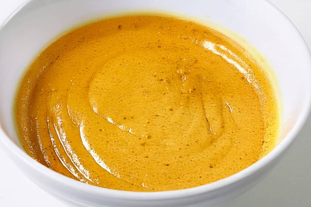 A white bowl filled with a creamy, golden-brown sauce, likely a curry or cheese-based dip, sits invitingly. The surface is smooth with slight swirls evident, suggesting it has been recently stirred. The background is plain, enhancing the focus on the vibrant color of the sauce that pairs perfectly with a slice of Pumpkin Swiss Roll.