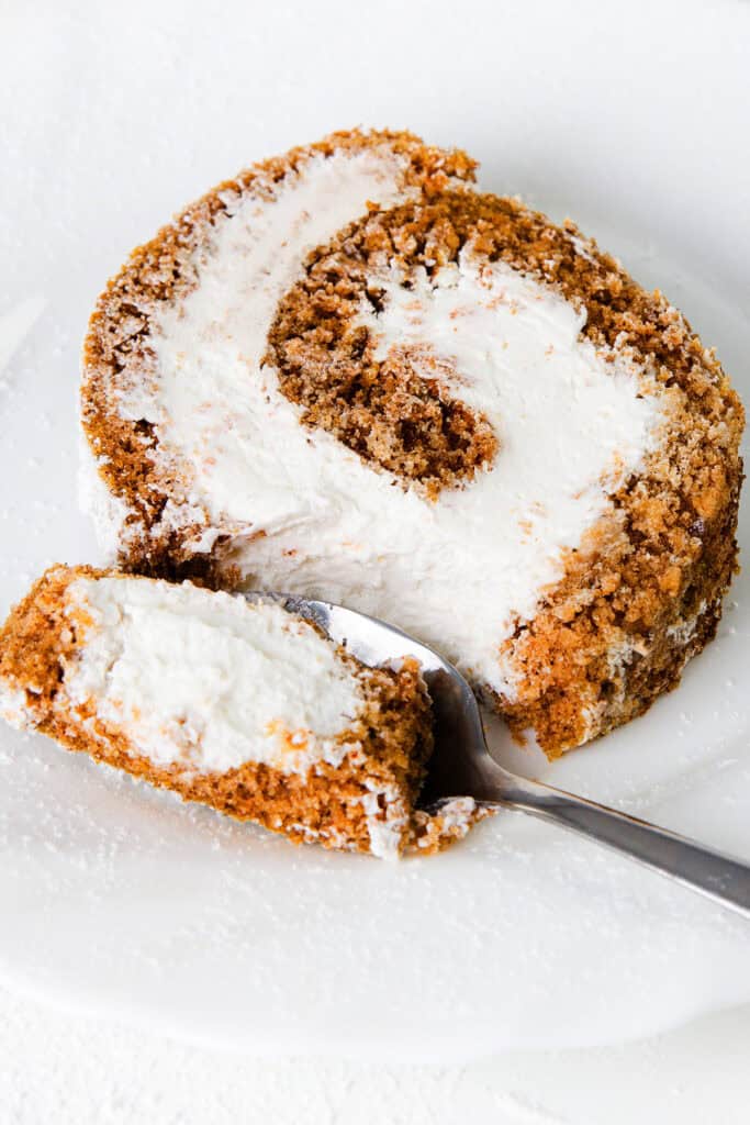 A close-up photo of a pumpkin Swiss roll slice with layers of light brown sponge cake and white cream filling, placed on a white plate. A spoon is next to the cake, with a small piece of the roll on it, ready to be eaten.