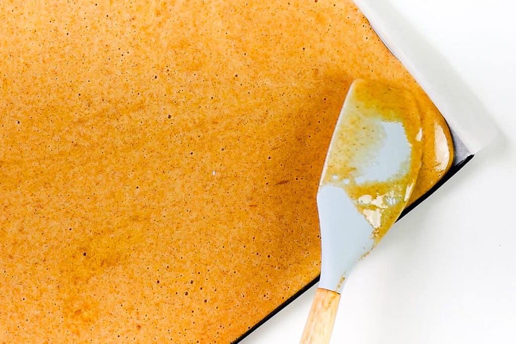 A silicone spatula spreads a thin, even layer of caramel-colored liquid on a white, parchment-lined baking sheet. The mixture appears smooth with small bubbles, suggesting it might be the beginning stages of a Pumpkin Swiss Roll or another sugary confection being prepared.